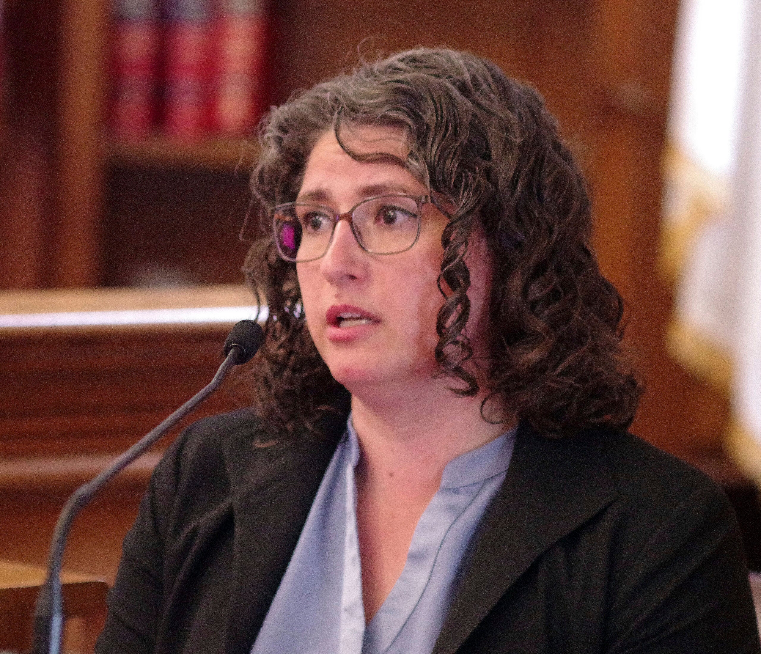 A witness with glasses speaking at a courtroom microphone.