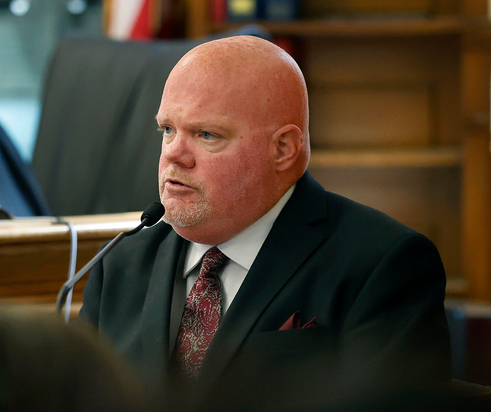 A witness with a reddish beard speaking into a courtroom microphone.