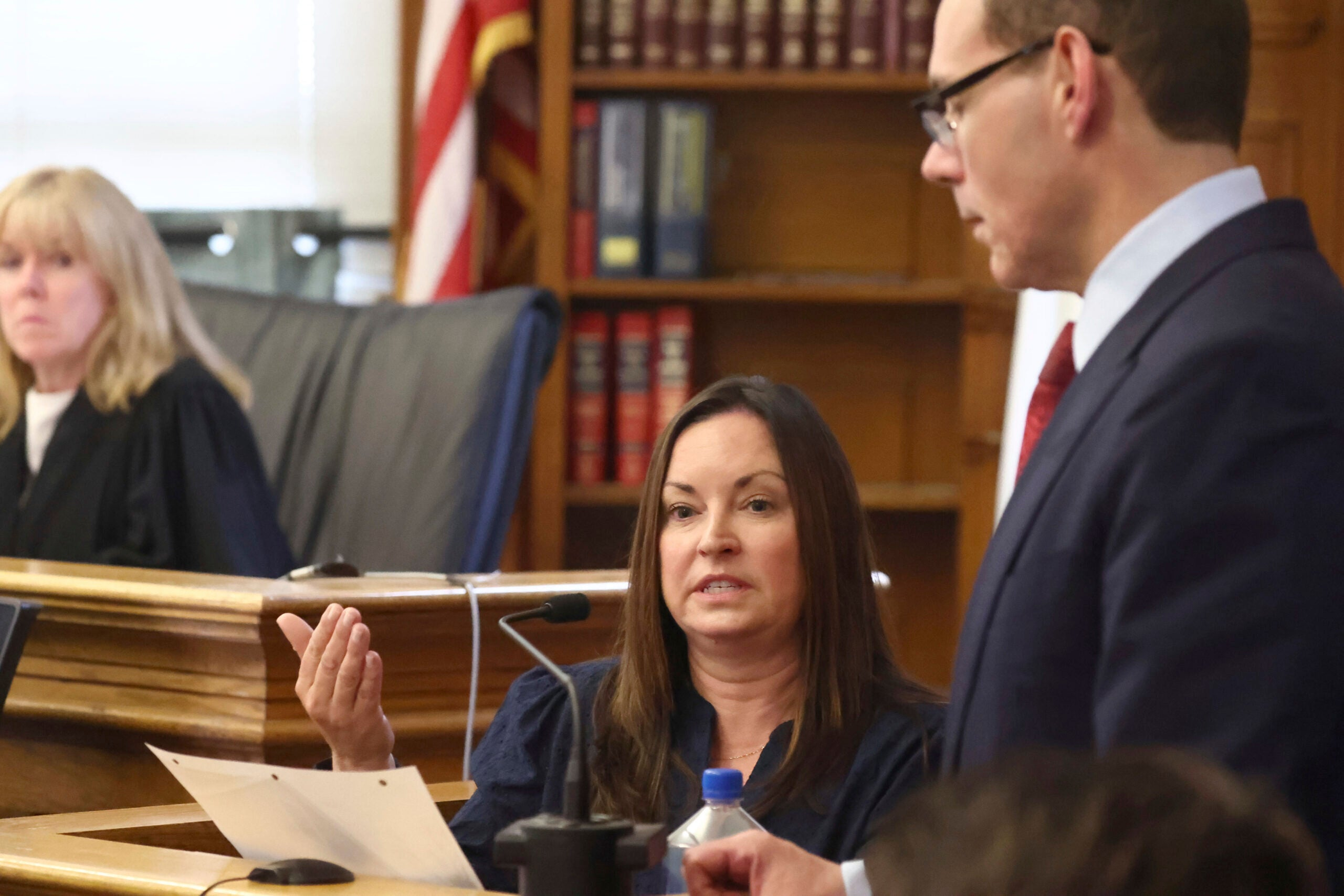 A witness speaking into a microphone with counsel standing beside the witness stand.