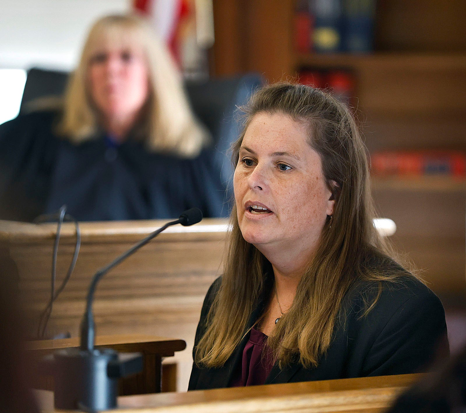 A witness with long hair at a courtroom microphone.