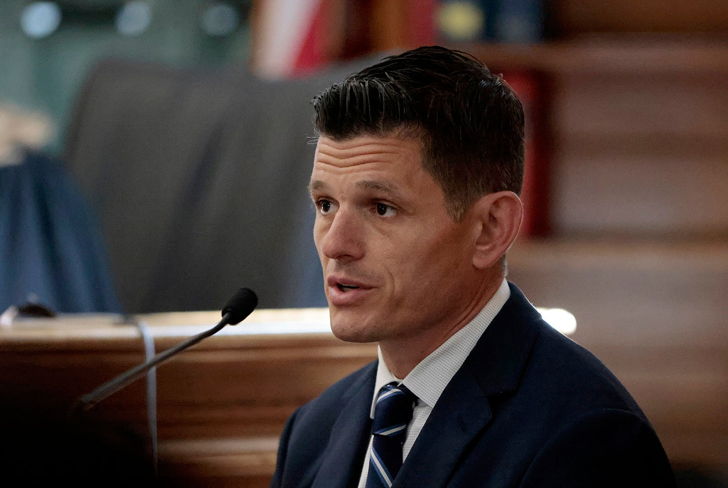 A witness in a dark suit and striped tie at a courtroom microphone.