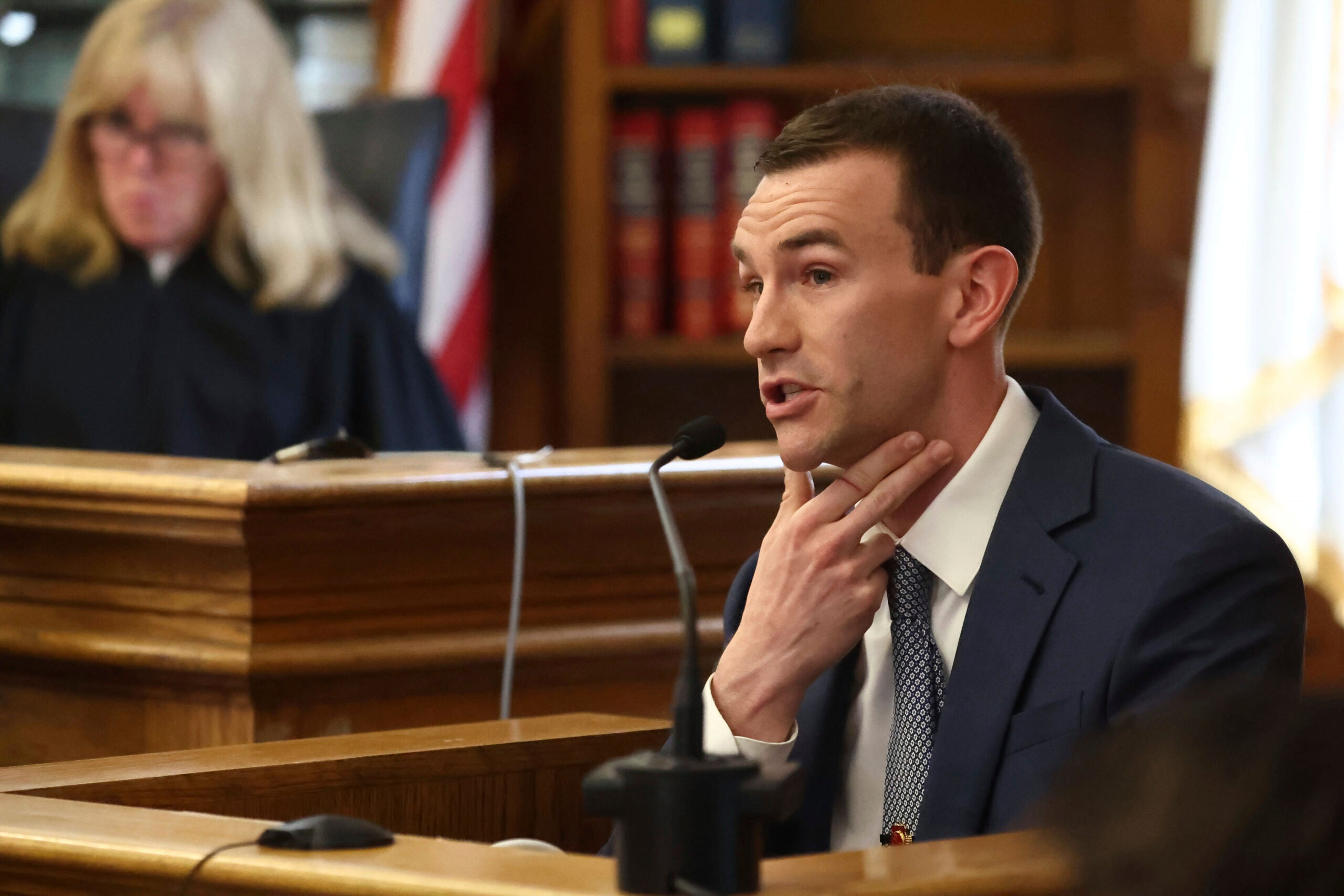 A witness places fingers against his neck while seated at a courtroom microphone.
