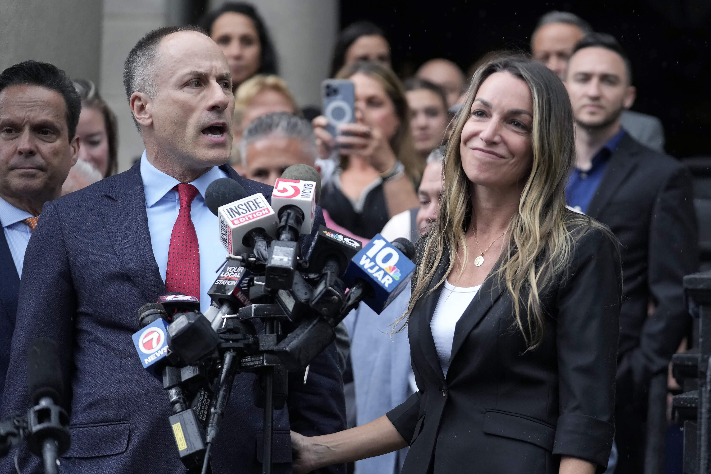 A press gathering outside court with microphones in front of a speaker and a woman beside him.